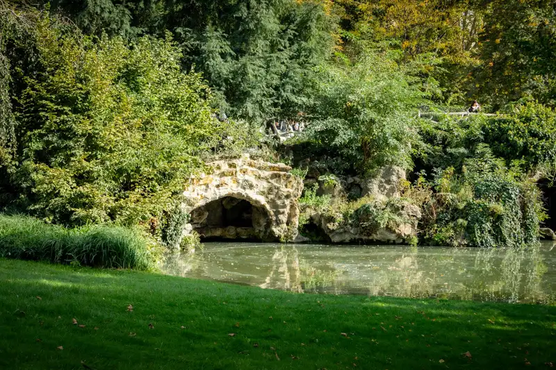 A tranquil park scene featuring dense greenery and a stone bridge reflecting on a peaceful river.