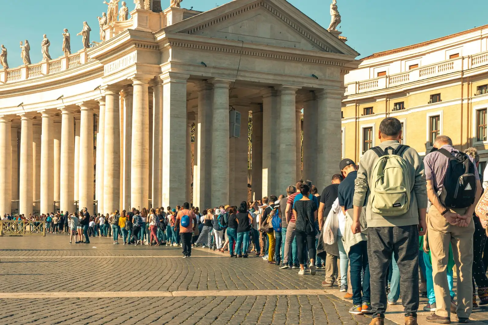 A large crowd of tourists lines up in sunny St. Peter's Square, showcasing architectural columns.